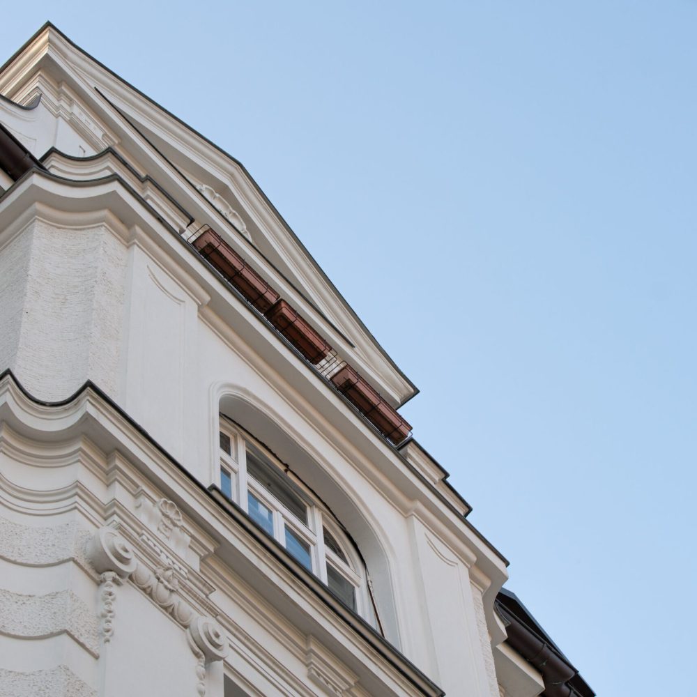Nahaufnahme einer verschnörkelten, weißen Gebäudefassade mit dekorativen architektonischen Details und einem Fenster vor einem klaren, blauen Himmel.