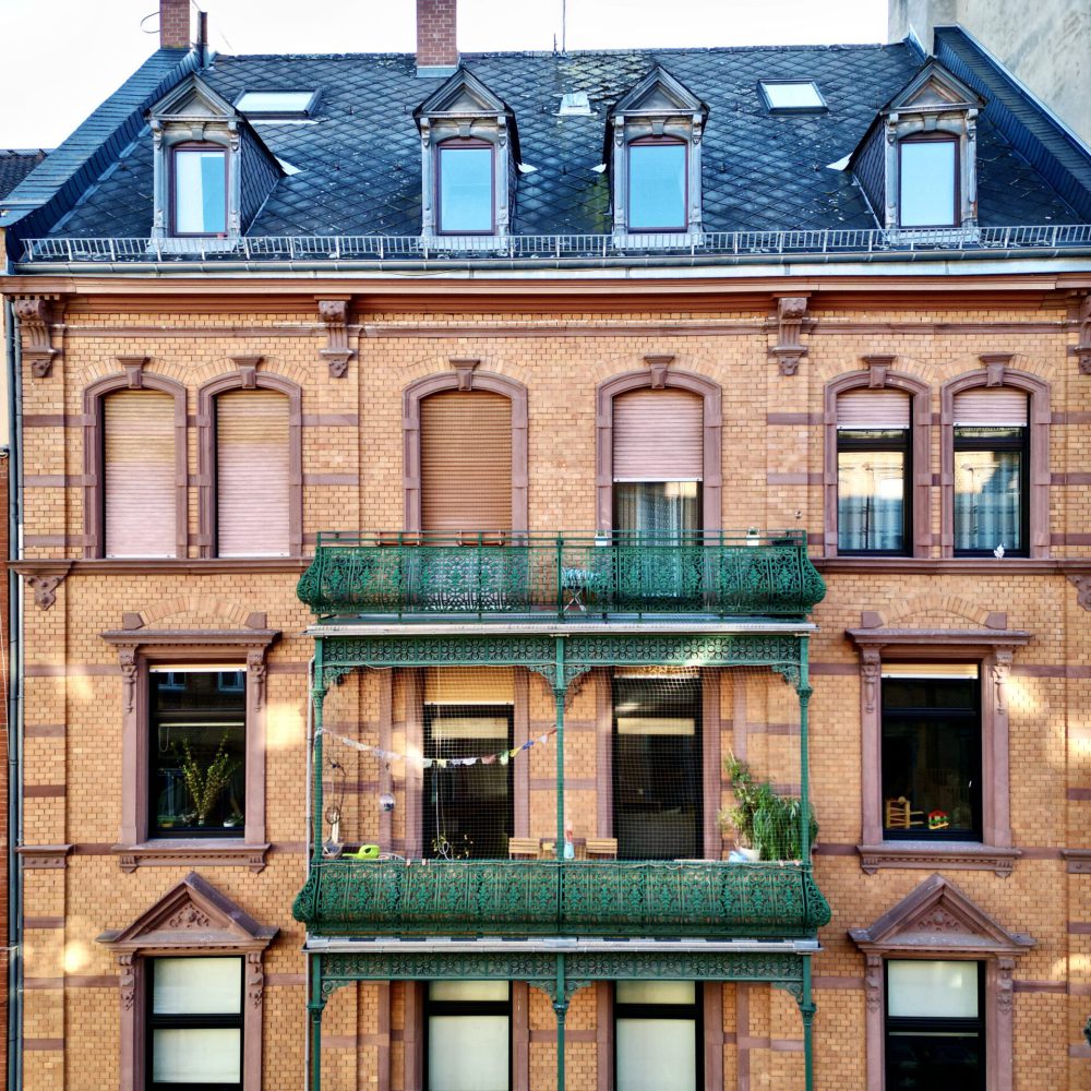 Ein roter Backsteinbau mit geschlossenen braunen Fensterläden, grünen Balkonen und dekorativen Verzierungen, der an einem sonnigen Tag zwischen zwei anderen städtischen Gebäuden liegt.
