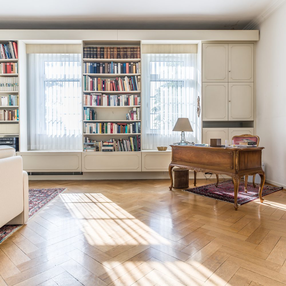Helles, geräumiges Wohnzimmer mit Holzfußboden, Bücherregalen voller Bücher, großen Fenstern mit transparenten Vorhängen, beigen Sesseln, einem Teppich und einem alten Holztisch mit Lampe und Stuhl.